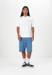 Hvid t-shirt med korte ærmer og logo, parret med lyseblå denimshorts. Sorte sneakers med hvide detaljer og hvide sokker.