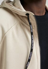Detailní záběr na béžovou mikinu na zip s černou šňůrkou, na které je bílé text "Active Wear Design", nošenou přes černé tričko s kulatým výstřihem.