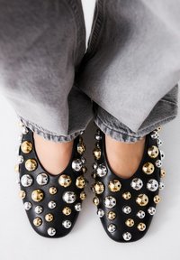 Zapatos negros tipo slip-on decorados con tachuelas de diferentes tamaños en oro y plata, con puntera redonda y textura suave. Jeans grises parcialmente visibles.