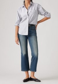 Chemise à rayures boutonnée blanche et marine, associée à un jean en denim bleu taille mi-haute. Des chaussures plates noires complètent la tenue.