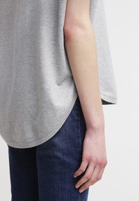 T-shirt grigia in cotone con orlo curvo, maniche corte e vestibilità rilassata, abbinata a jeans blu scuro. Pelle visibile sul braccio.