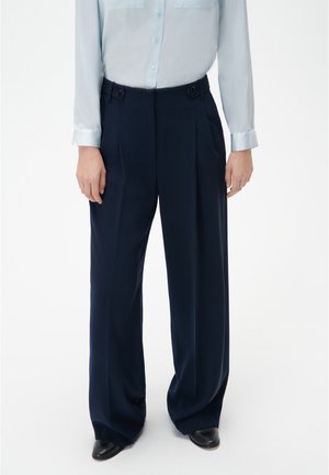 Navy pleated trousers made of smooth fabric with a high waist, featuring button accents and wide legs. Paired with a light blue shirt.