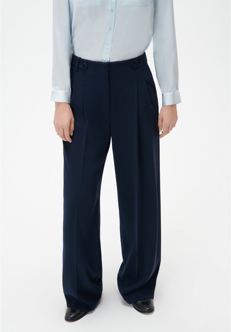 Pantalons plissés navy en tissu lisse avec une taille haute, dotés d'accents de boutons et de jambes larges. Associés à une chemise bleu clair.