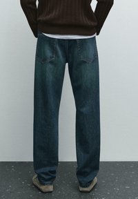 Jeans in denim blu scuro con vestibilità ampia, caratterizzati da un classico design a cinque tasche e una leggera scoloritura, abbinati a un maglione marrone a coste.