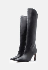 Bottes hautes en cuir noir jusqu'au genou, avec un bout pointu, un talon aiguille et une texture lisse. Détail de fermeture éclair latérale visible.