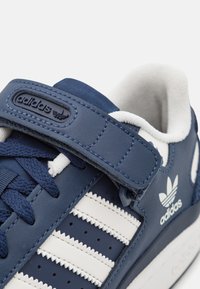 Blå lædersneakers med hvide striber, udstyret med en Velcro-rem, teksturerede detaljer og et logopatch på siden.