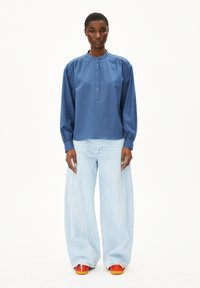 Donna in piedi di fronte, che indossa una camicia blu con bottoni, jeans larghi di un blu chiaro e sneaker arancioni, su uno sfondo bianco.