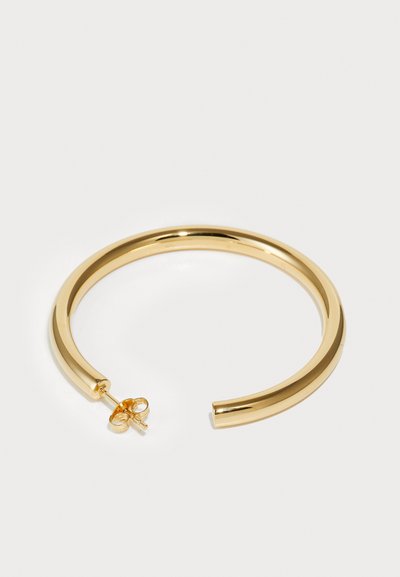 Pulsera de manga de metal en tono dorado con un acabado liso y pulido. Presenta un diseño abierto y un pequeño broche para un cierre seguro.