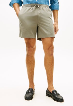 Mężczyzna w beżowych krótkich spodenkach ze ściągaczem, czarnych mokasynach i podwiniętej długiej koszuli dżinsowej, stojący z rękami w kieszeniach.