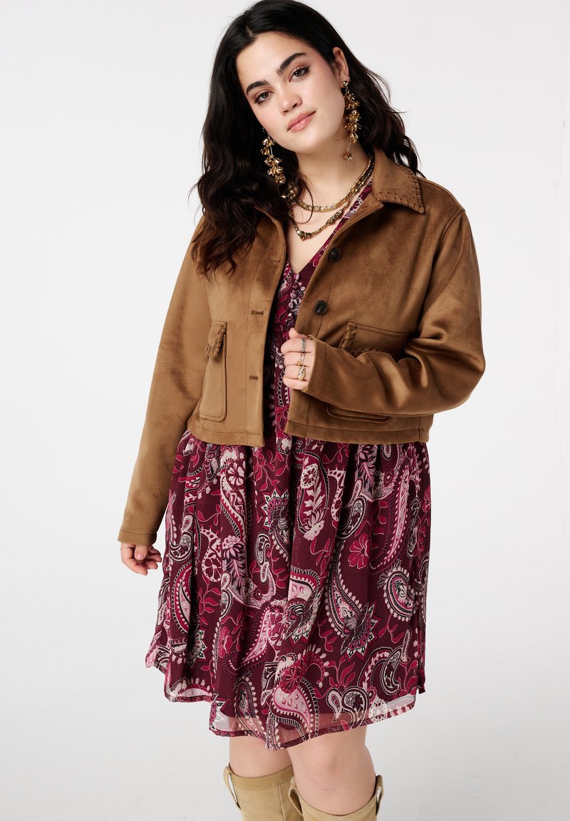 Veste courte en suède marron avec deux poches frontales, associée à une robe bordeaux à motif cachemire, dotée d'une jupe évasée et d'un tissu superposé.