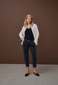 Moteris su blondinais plaukais dėvi baltą sagotu marškinių viršų, juodą palaidinę, tamsiai mėlynas trumpintas kelnes, juodą diržą ir juodus aukštakulnius, stovinčią ant pilkos grindų.