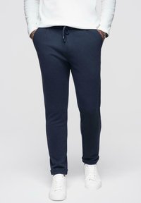 Homme portant un pantalon de jogging bleu marine à cordon de serrage, des baskets blanches et un haut blanc à manches longues, les mains dans les poches.