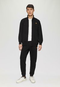 Schwarze Zip-Jacke mit Stehkragen und Logo, kombiniert mit passenden schwarzen Jogginghosen. Beide aus einem weichen Stoff. Weiße Sneakers mit schwarzen Akzenten.