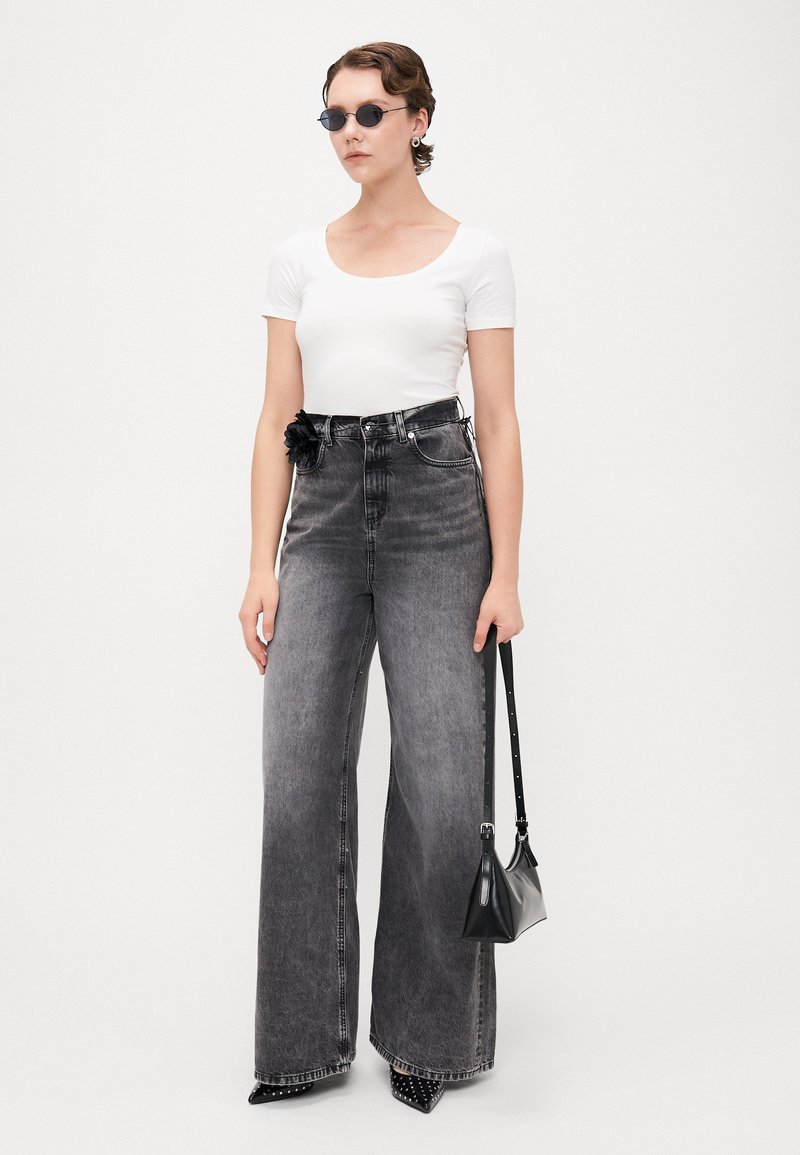 Jeans grigi alti in vita con gambe larghe e un dettaglio floreale in vita, abbinati a una blusa bianca aderente e occhiali da sole neri. Borsa nera.