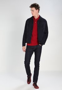 Navy suede jacket over a red polo, dark denim jeans, and burgundy sneakers. Features two chest pockets and a straight fit. Clean design.