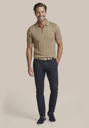 Smiling man wearing a beige short-sleeve polo shirt, dark blue pants, light woven belt, and navy slip-on shoes standing with one hand in pocket.