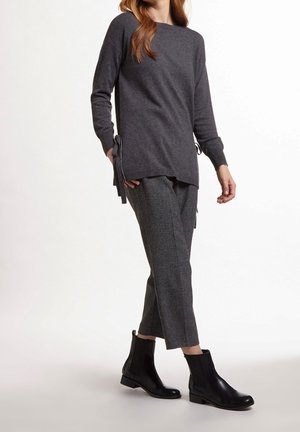 Frau trägt einen dunkelgrauen Pullover mit seitlichen Bindebändern, graue karierte kurze Hosen und schwarze Stiefeletten vor weißem Hintergrund.