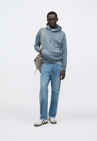 Grå hættetrøje lavet af blødt stof, kombineret med lyseblå denimjeans. Modellen har grå sneakers med sorte striber og bærer en tanfarvet taske.