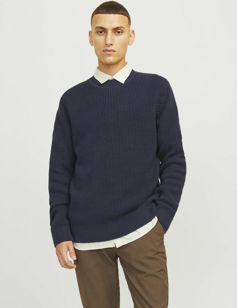 Marinblå stickad tröja med ett strukturerat mönster, rund halsringning och ribbade ärmslut, över en ljusrandig skjorta och bruna byxor.