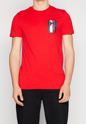 Homme portant un t-shirt rouge à manches courtes avec un logo noir, blanc et rouge sur la poitrine gauche, assorti à un pantalon noir.