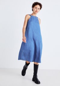 Lys blå denimkjole med en løs A-linjeform, som har en rynket halslinje og tynne stropper. Kombinert med støvletter som går til kneet i svart.