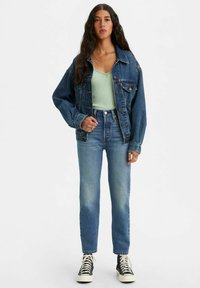 Denim jacket in dark blue, paired with a light green fitted top and high-waisted blue jeans. Black high-top sneakers complete the look.