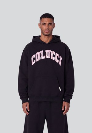 Uomo che indossa una felpa nera con cappuccio con la scritta "COLUCCI" stampata in grandi lettere rosa chiaro sul petto, in piedi contro uno sfondo semplice.
