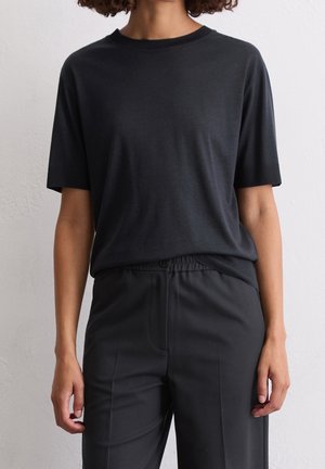 Vrouw draagt een effen donker T-shirt met korte mouwen, ingestopt in een donkere broek met hoge taille, staand tegen een lichte gestructureerde muur.