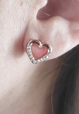 Herzförmige Ohrringe aus roségold mit einem klaren Kristallrand und einem inneren pinken Element, die auf einem hellen Hautton liegen.