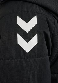 Veste matelassée noire en tissu avec un design en chevron blanc sur le bras gauche. Le matériau a une finition texturée avec des coutures visibles.