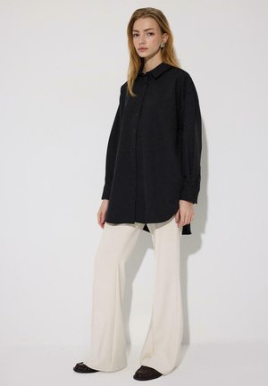 Kobieta ubrana w długą czarną koszulę na guziki, kremowe spodnie o szerokich nogawkach i ciemne buty, stojąca na tle jasnego, prostego tła.