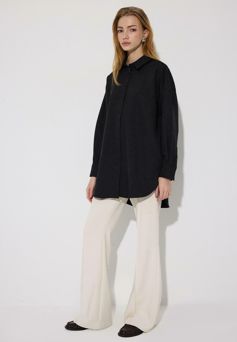 Γυναίκα που φοράει μακρύ μαύρο πουκάμισο με κουμπιά, κρεμ παντελόνι με φαρδιά μπατζάκια και σκούρα παπούτσια, στέκεται μπροστά σε απλό φωτεινό φόντο.