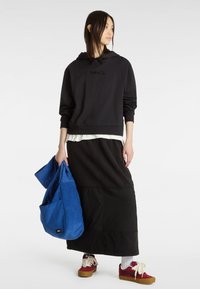 Felpa nera con cappuccio e logo ricamato, sovrapposta a una camicia bianca. Gonna nera lunga, borsa blu e sneakers rosse con lacci bianchi.