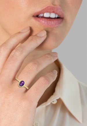 Guldring med en oval lilla ædelsten, båret på en finger. Hånden rører ved hagen, mod en neutral baggrund med en krave synlig.