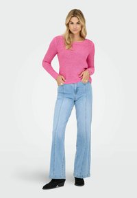 Femme debout les mains dans les poches, portant un pull rose à manches longues, un jean taille haute bleu clair et des bottines noires.