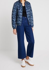 Gequiltede marineblauwe pufferjack met bloemenpatroon, gecombineerd met hoge taille, wijde denims in donker denim en witte sneakers met beige zolen.