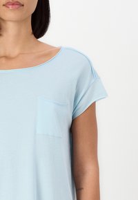 Světle modrý krátký rukáv top vyrobený z měkké látky, s kulatým výstřihem a malou kapsičkou na prsou. Minimalistický design s hladkou strukturou.