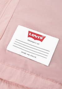 Ανοιχτό ροζ ύφασμα με ξεχωριστή υφή, που φέρει μια λευκή ετικέτα με το κόκκινο λογότυπο της Levi's και γραμμές για εξατομίκευση. Το κείμενο λέει "HAND-ME-DOWN."