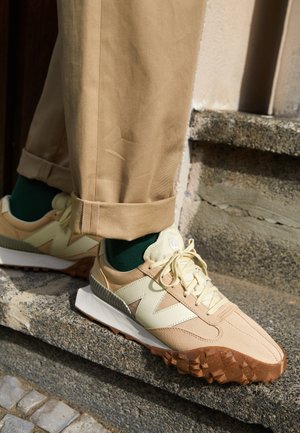 Beżowe i kremowe sneakersy z grubą, teksturowaną brązową podeszwą, noszone z beżowymi spodniami podwiniętymi u dołu i ciemnozielonymi skarpetkami, stojące na kamiennym stopniu.