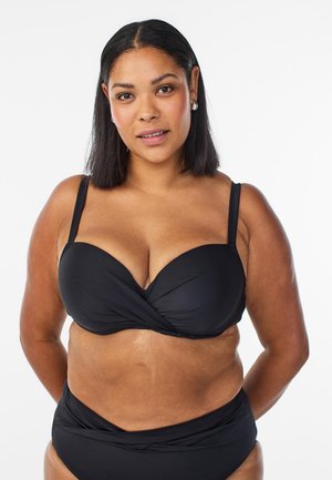 Femme aux cheveux raides et foncés portant un bikini noir avec des détails torsadés, debout devant un fond blanc uni.