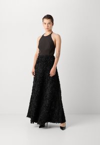 Haut halter noir associé à une longue jupe noire texturée avec des détails en appliqué floral. Des chaussures à talons hauts noires classiques complètent la tenue.