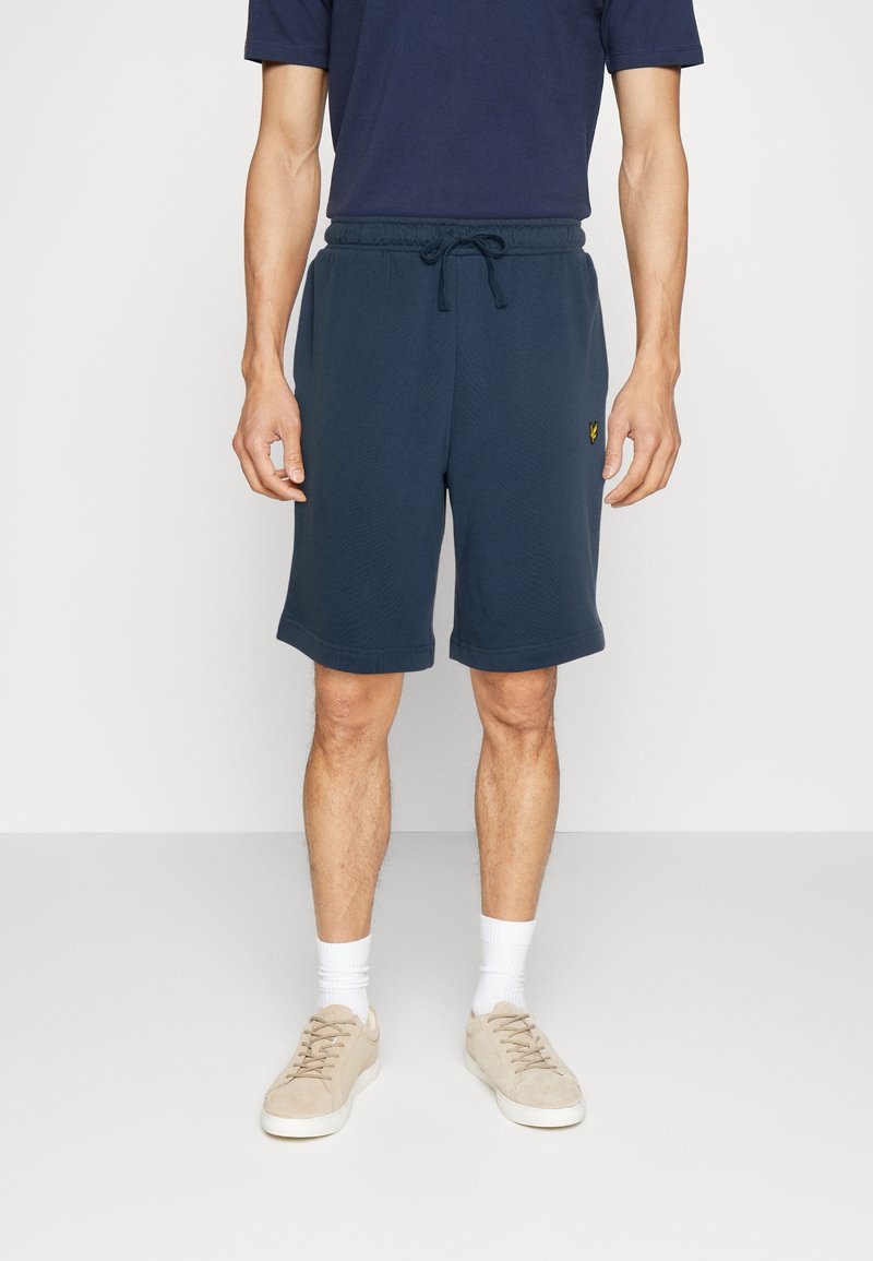 Shorts bleus marine avec un motif texturé, taille élastique et cordon de serrage. Présente un logo dans le coin inférieur gauche. Portés avec des chaussures claires.