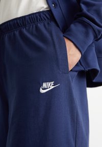 Temno modri Nike trening hlače z elastiko v pasu. Imajo bel Nike logo na levem stegnu in gladko, teksturirano tkanino.