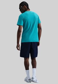 Camiseta de manga corta color turquesa, pantalones cortos cargo de color marino, calcetines deportivos blancos, zapatillas ligeras con detalles de acento, vistas desde una perspectiva trasera.
