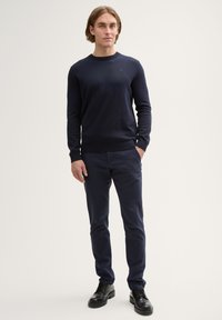 Jeune homme debout portant un pull bleu marine, un pantalon bleu marine assorti et des chaussures de ville noires sur un fond clair uni.
