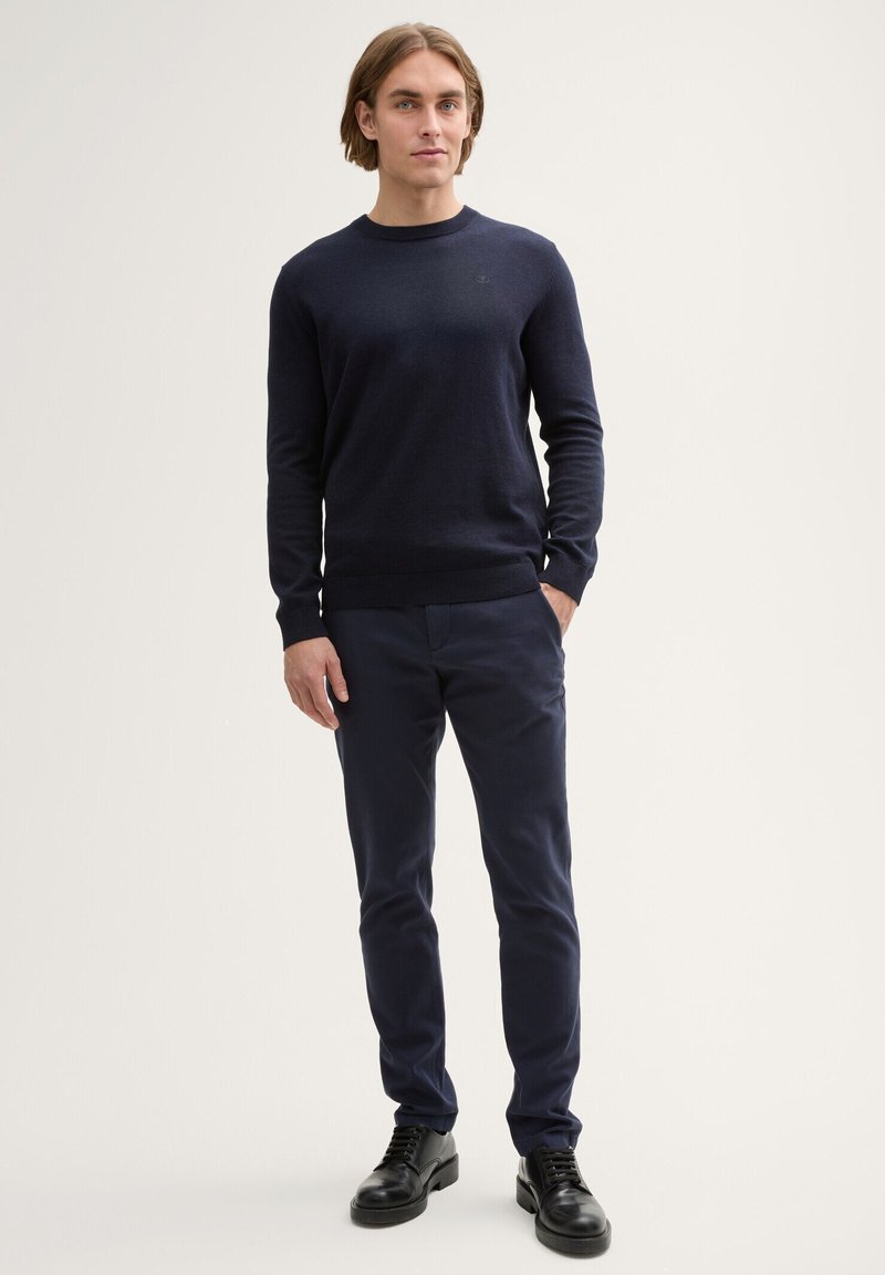 Jeune homme debout portant un pull bleu marine, un pantalon bleu marine assorti et des chaussures de ville noires sur un fond clair uni.