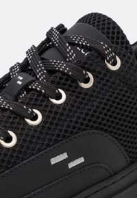 Černá sportovní obuv s horní částí z prodyšné síťoviny, kulatou špičkou, stříbrnými průchodkami, texturovanými detaily a kontrastními bílými prvky na podrážce.