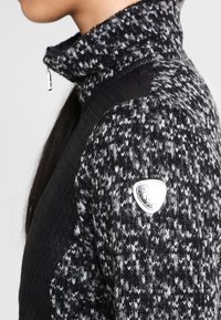 Svart och gråmönstrad tröja med hög krage, med dragkedjestängning och en silverfärgad logobadge på vänster axel. Texturerad tyg.
