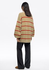 Długi, oversize sweter w kolorze beżowym z czerwonymi paskami, wykonany z miękkiej, teksturowanej tkaniny; ma opadające ramiona i okrągły dół. W zestawie z ciemnymi spodniami typu wide-leg.