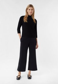 Schwarzer Pullover mit hohem Kragen, kombiniert mit weit geschnittenen schwarzen Hosen und schwarzen Schuhen. Das Ensemble zeichnet sich durch klare Linien und eine glatte Textur aus.